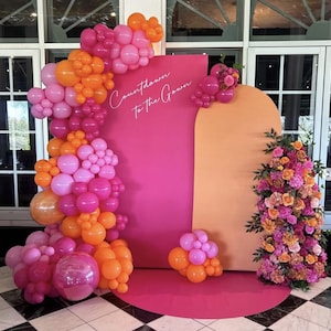 May include: A vibrant event backdrop featuring a pink and orange color scheme. A large pink rectangular panel displays the text "Countdown to the Gown" in white script. The scene is decorated with balloon clusters in shades of pink and orange, and a floral arrangement.