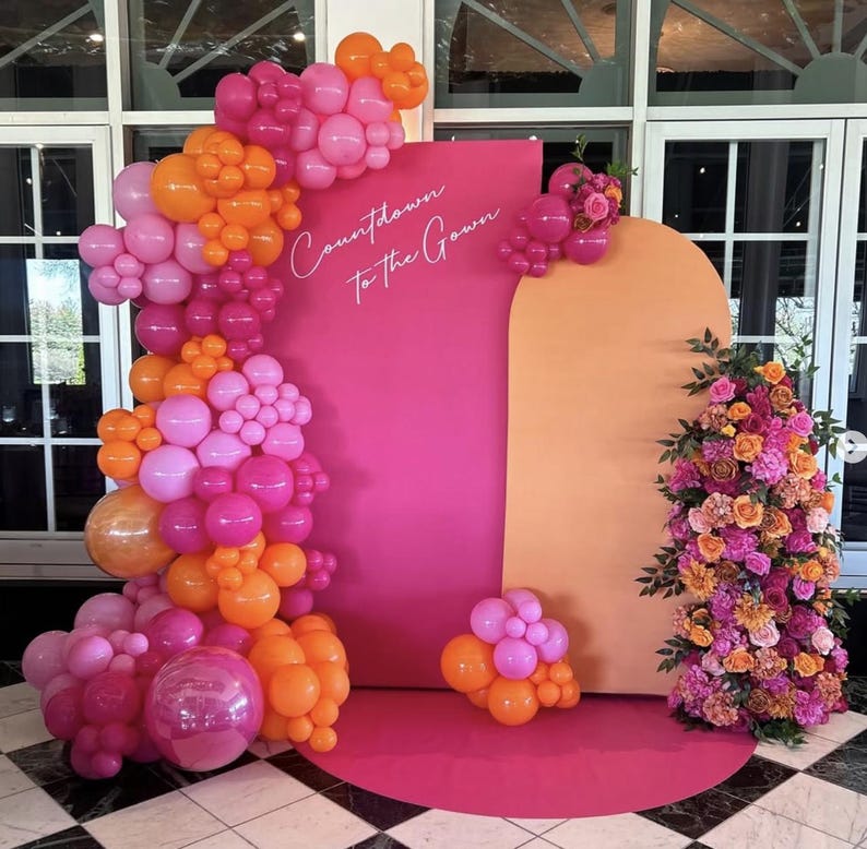 May include: A vibrant event backdrop featuring a pink and orange color scheme. A large pink rectangular panel displays the text "Countdown to the Gown" in white script. The scene is decorated with balloon clusters in shades of pink and orange, and a floral arrangement.