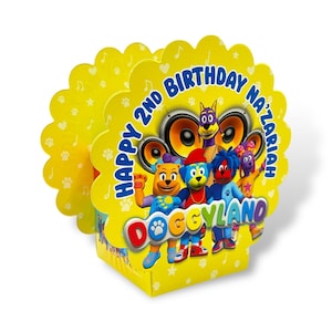 May include: Yellow and blue birthday party favor box with a cartoon dog character on the front. The box says "Happy 2nd Birthday Nazariah Doggyland".