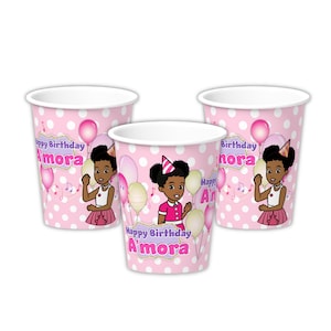 May include: Set of three pink paper cups with a white polka dot pattern. Each cup features a cartoon illustration of a young girl with brown skin and curly hair wearing a pink dress and a party hat. The cups have the text "Happy Birthday Amora" printed on them.