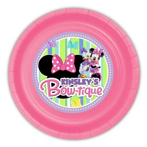 Puede incluir: Un plato de papel rosa con Minnie Mouse y Daisy Duck. El plato tiene el texto "Kinsley's Bow-tique" y una silueta de Minnie Mouse con un lazo rosa de lunares. El fondo tiene rayas verdes y azules.