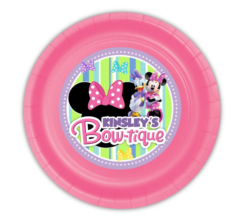 Puede incluir: Un plato de papel rosa con Minnie Mouse y Daisy Duck. El plato tiene el texto "Kinsley's Bow-tique" y una silueta de Minnie Mouse con un lazo rosa de lunares. El fondo tiene rayas verdes y azules.