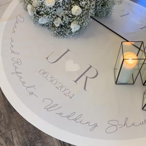 May include: A round white wedding shower decoration with floral arrangements and a lit candle. The decoration features the initials "J R", a heart, and the date "06.30.2024". The words "Dear and Rafael's Wedding Shower" are also written on the decoration.