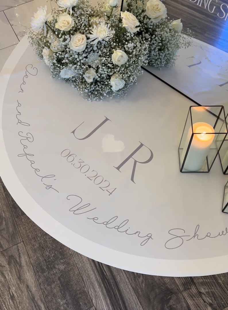 May include: A round white wedding shower decoration with floral arrangements and a lit candle. The decoration features the initials "J R", a heart, and the date "06.30.2024". The words "Dear and Rafael's Wedding Shower" are also written on the decoration.