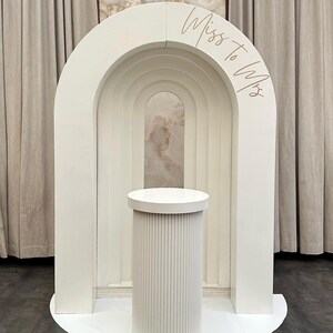 May include: A white wedding arch with a cylindrical pedestal. The arch has a layered design with the text "Miss to Mrs" in a gold script. The pedestal has vertical ribbed detailing and a circular top.