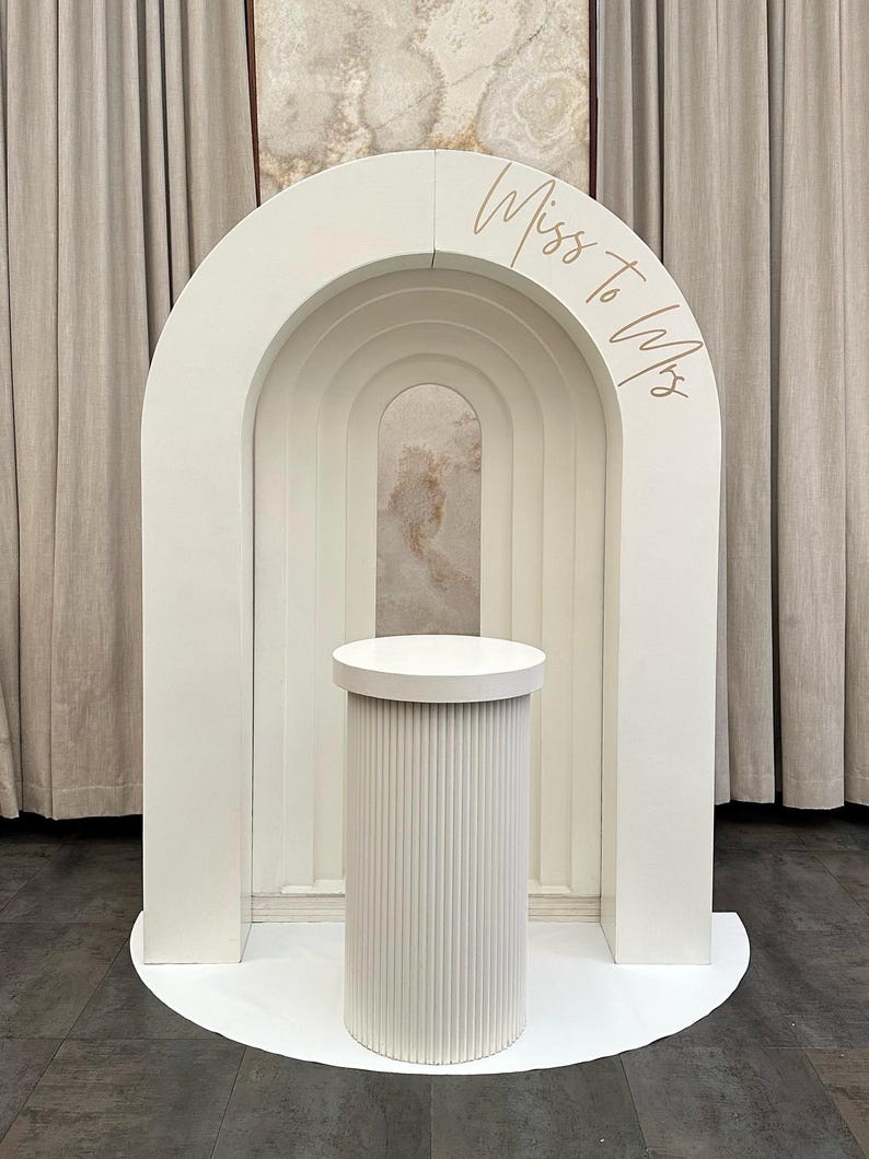 May include: A white wedding arch with a cylindrical pedestal. The arch has a layered design with the text "Miss to Mrs" in a gold script. The pedestal has vertical ribbed detailing and a circular top.
