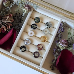 以下が含まれることがあります： 金色のフレームのガラス製ジュエリーボックスに飾られた、透明な樹脂に封入されたドライフラワーのハンドメイドリングのコレクション。リングは様々な花の模様が特徴で、シルバーとゴールドのバンドにセットされています。ボックスの周りにはドライフラワーと花びらが飾られています。