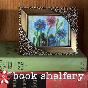 May include: A small framed artwork featuring a floral painting with blue and purple flowers. The frame is ornate, with a gold finish. The artwork rests on a stack of books, including "Sunset Pass" by Zane Grey. The text "the book shelfery" is visible.