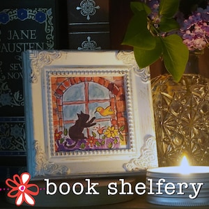 May include: A framed watercolor painting depicts a black cat reaching for a yellow bird outside a window. The white frame has ornate detailing. A lit candle, a vase of purple flowers, and books are in the background. The text "book shelfery" is at the bottom.
