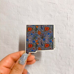 Puede incluir: Una pegatina del estado de Nuevo M&eacute;xico con un patr&oacute;n floral en tonos azules, naranjas y verdes. La pegatina tiene un borde blanco y el texto "@michellelacreations" en la esquina inferior derecha.