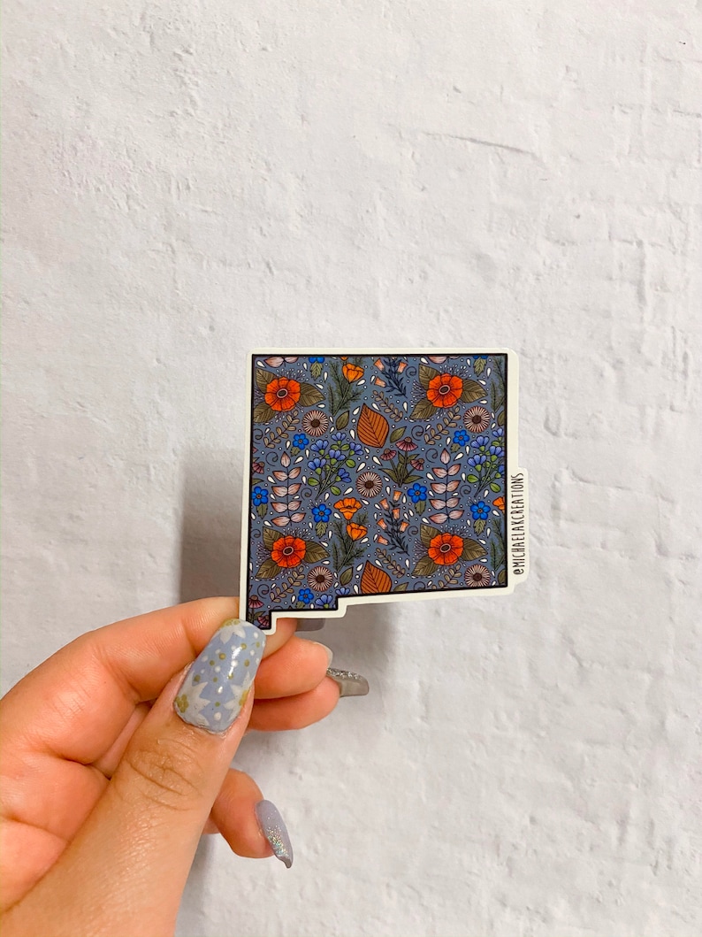 Puede incluir: Una pegatina del estado de Nuevo M&eacute;xico con un patr&oacute;n floral en tonos azules, naranjas y verdes. La pegatina tiene un borde blanco y el texto "@michellelacreations" en la esquina inferior derecha.