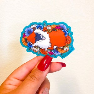 Puede incluir: Una pegatina con un conejillo de indias de dibujos animados rodeado de flores coloridas. El conejillo de indias es naranja y blanco con ojos negros y una nariz rosa. Las flores son de varios colores, incluyendo rojo, amarillo, naranja, azul y morado. La pegatina est&aacute; sobre un fondo turquesa.