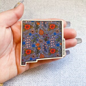 Puede incluir: Una pegatina del estado de Nuevo M&eacute;xico con un patr&oacute;n floral en tonos naranja, azul y verde sobre un fondo morado. La pegatina tiene un borde blanco y el texto "@michaelakcreations" en la esquina inferior derecha.