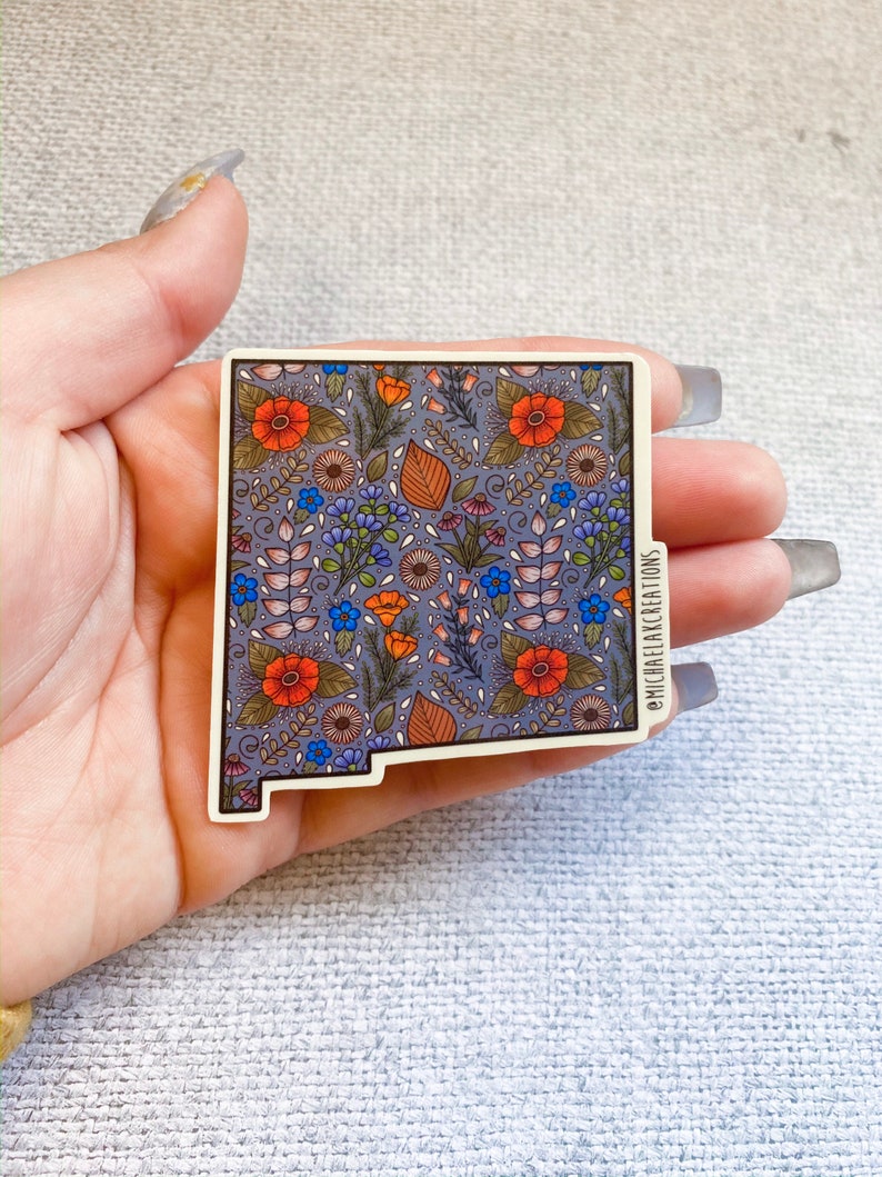 Puede incluir: Una pegatina del estado de Nuevo M&eacute;xico con un patr&oacute;n floral en tonos naranja, azul y verde sobre un fondo morado. La pegatina tiene un borde blanco y el texto "@michaelakcreations" en la esquina inferior derecha.