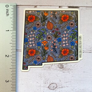 Puede incluir: Una pegatina con un patr&oacute;n floral en tonos naranja, azul y verde sobre un fondo gris. La pegatina tiene la forma del estado de Nuevo M&eacute;xico e incluye el texto "@MICHAELAKCREATIONS".