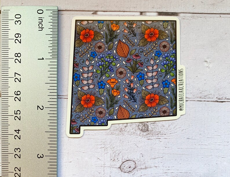 Puede incluir: Una pegatina con un patr&oacute;n floral en tonos naranja, azul y verde sobre un fondo gris. La pegatina tiene la forma del estado de Nuevo M&eacute;xico e incluye el texto "@MICHAELAKCREATIONS".