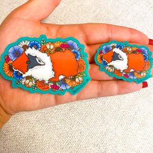 Puede incluir: Dos pegatinas coloridas con un conejillo de indias de dibujos animados con un dise&ntilde;o floral. El conejillo de indias es naranja, blanco y negro. Las pegatinas est&aacute;n en una mano.