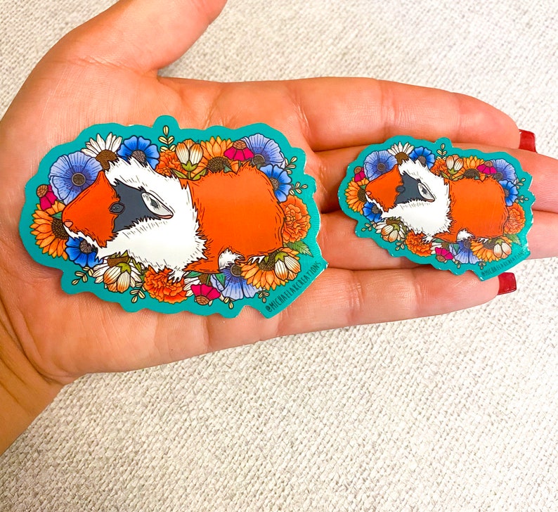 Puede incluir: Dos pegatinas coloridas con un conejillo de indias de dibujos animados con un dise&ntilde;o floral. El conejillo de indias es naranja, blanco y negro. Las pegatinas est&aacute;n en una mano.
