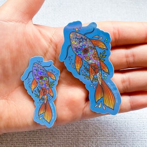 K&ouml;nnte beinhalten: Zwei schimmernde holografische Aufkleber mit Koi-Fischen und floralen Designs auf blauem Hintergrund. Der gr&ouml;&szlig;ere Aufkleber tr&auml;gt den Text "@michelle.ray.creations" in der unteren rechten Ecke.