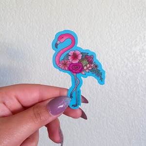 May include: A pink flamingo sticker with a blue background. The flamingo is decorated with pink, red, and green flowers. The sticker is on a white background.