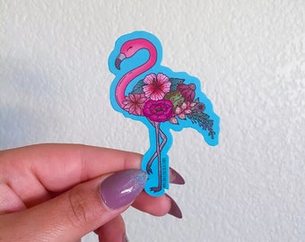 May include: A pink flamingo sticker with a blue background. The flamingo is decorated with pink, red, and green flowers. The sticker is on a white background.