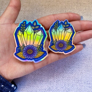 May include: Two iridescent holographic stickers featuring a cluster of yellow crystals with blue flowers and green leaves. The stickers have the text "@michaelakcreations" on them.