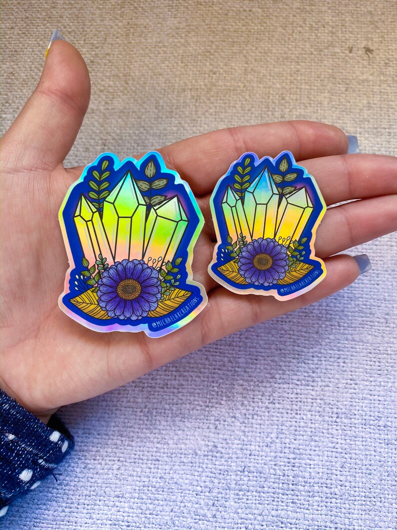 May include: Two iridescent holographic stickers featuring a cluster of yellow crystals with blue flowers and green leaves. The stickers have the text "@michaelakcreations" on them.