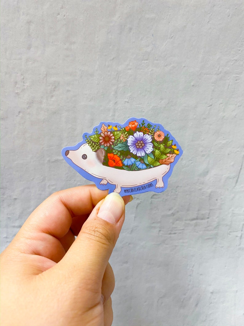 May include: A sticker featuring a cartoon hedgehog with a floral arrangement on its back. The hedgehog is white with a light blue outline, and the flowers are a mix of orange, purple, and yellow. The sticker has the text "@MICHAELAKREATIONS".