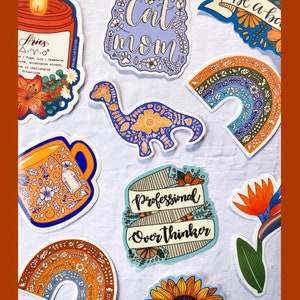 K&ouml;nnte beinhalten: Eine Sammlung bunter Aufkleber mit verschiedenen Designs, darunter eine Kerze, eine Tasse, ein Dinosaurier, ein Regenbogen, eine Sonnenblume und ein Banner mit der Aufschrift "Professional Overthinker". Die Aufkleber befinden sich alle auf einem wei&szlig;en Hintergrund. Der Text "NEW DESIGNS CREATED" befindet sich oben im Bild, und der Text "EVERY MONTH" befindet sich unten.