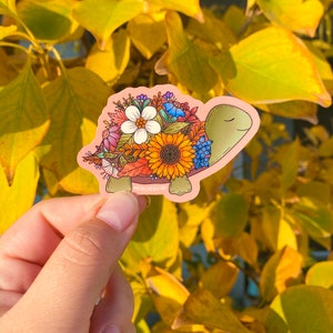 May include: A sticker of a turtle with a shell made of colorful flowers, including sunflowers, bluebells, and other wildflowers. The turtle is on a pink background.