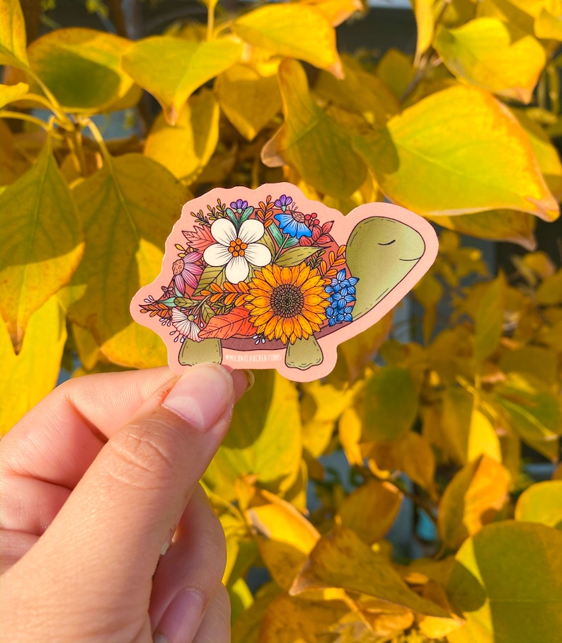 May include: A sticker of a turtle with a shell made of colorful flowers, including sunflowers, bluebells, and other wildflowers. The turtle is on a pink background.