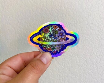 May include: A holographic sticker featuring a planet with a ring, filled with colorful flowers and greenery. The sticker has a blue border and a rainbow iridescent background.