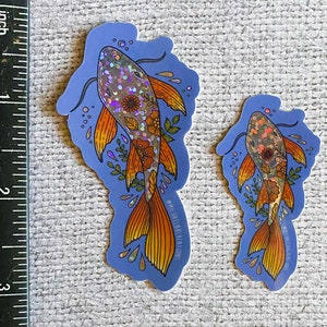 K&ouml;nnte beinhalten: Zwei schimmernde holografische Aufkleber mit Koi-Fischen und floralen Details auf blauem Hintergrund. Der gr&ouml;&szlig;ere Aufkleber ist etwa 7,6 cm hoch und der kleinere etwa 5 cm hoch. Beide Aufkleber haben den Text "MICHAEL ELIAS CREATIONS" unten.