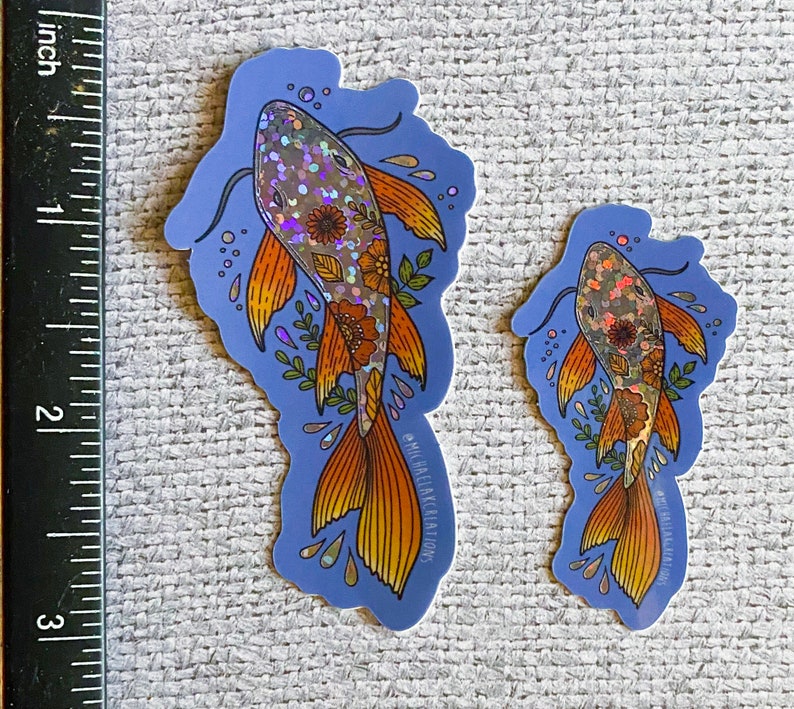K&ouml;nnte beinhalten: Zwei schimmernde holografische Aufkleber mit Koi-Fischen und floralen Details auf blauem Hintergrund. Der gr&ouml;&szlig;ere Aufkleber ist etwa 7,6 cm hoch und der kleinere etwa 5 cm hoch. Beide Aufkleber haben den Text "MICHAEL ELIAS CREATIONS" unten.