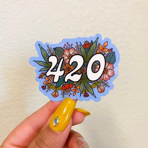 May include: A blue sticker with a floral design and the number 420 in white. The sticker is being held by a hand with yellow nail polish.