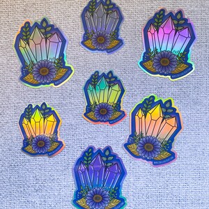 May include: Seven iridescent holographic stickers featuring a cluster of crystals with a flower and leaves. The crystals are in various shades of blue, purple, pink, and yellow. The stickers have a blue outline and the text "#MICHAELLABCREATIONS" is printed on each sticker.