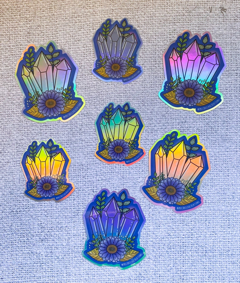 May include: Seven iridescent holographic stickers featuring a cluster of crystals with a flower and leaves. The crystals are in various shades of blue, purple, pink, and yellow. The stickers have a blue outline and the text "#MICHAELLABCREATIONS" is printed on each sticker.