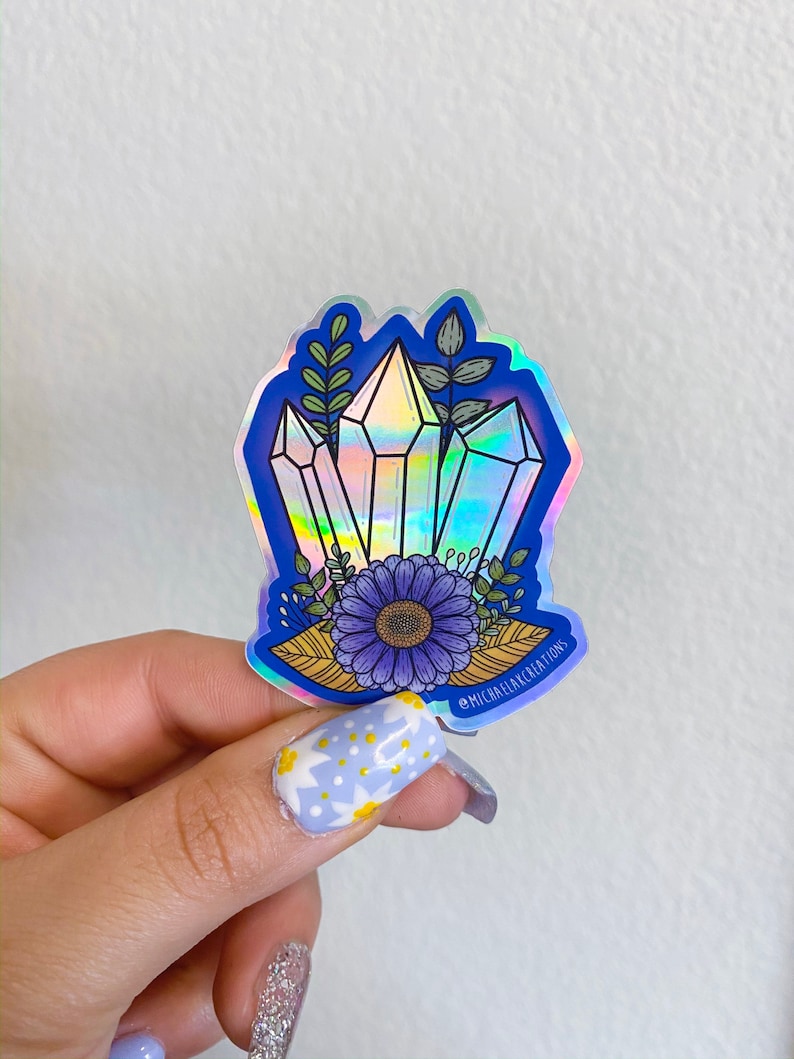 May include: A holographic sticker featuring a cluster of iridescent crystals with a purple flower and green leaves. The sticker has a blue background and the text "@michaelakcreations" in the bottom right corner.