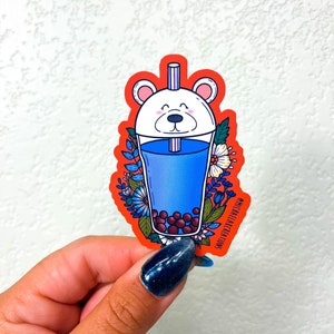 May include: A red sticker with a white polar bear face on a blue bubble tea cup with boba pearls. The cup is surrounded by flowers and leaves. The sticker says "#michaelscakacreations" at the bottom.