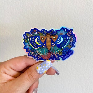 May include: A holographic sticker featuring a blue and green moth with floral details and crescent moons. The sticker has a white border and the text "@michaelflake.creations" in the bottom right corner.