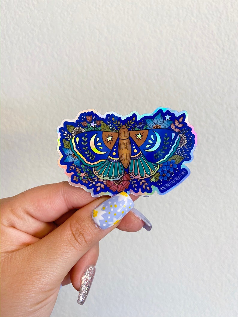 May include: A holographic sticker featuring a blue and green moth with floral details and crescent moons. The sticker has a white border and the text "@michaelflake.creations" in the bottom right corner.