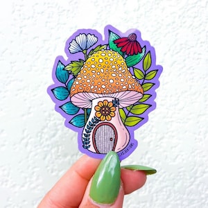 May include: A purple sticker with a whimsical mushroom house illustration. The mushroom is yellow and orange with white spots and a pink base. The house has a small door and a flower window. The mushroom is surrounded by green leaves and flowers.