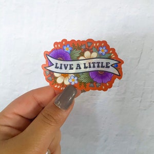 May include: A sticker with a floral design and the text "Live a Little" in a banner. The flowers are purple, white, and blue.