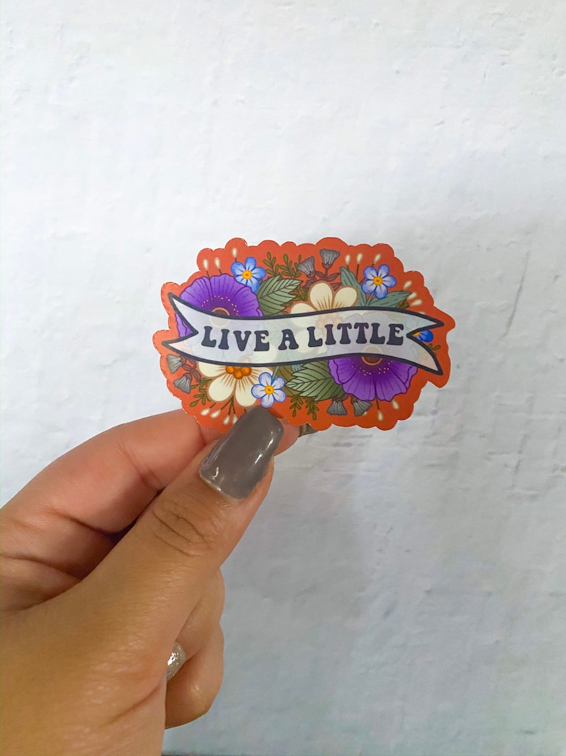 May include: A sticker with a floral design and the text "Live a Little" in a banner. The flowers are purple, white, and blue.