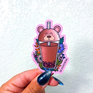 May include: Pink sticker with a cartoon illustration of a bear-shaped boba tea cup with a straw, surrounded by flowers. The text "MICHAELAELACREATIONS" is on the sticker.