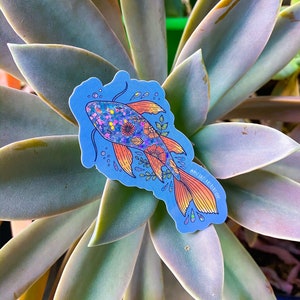 K&ouml;nnte beinhalten: Ein blauer und orangefarbener Fisch-Aufkleber mit Glitzer und floralen Details. Der Aufkleber befindet sich auf einer gr&uuml;nen Sukkulentenpflanze.