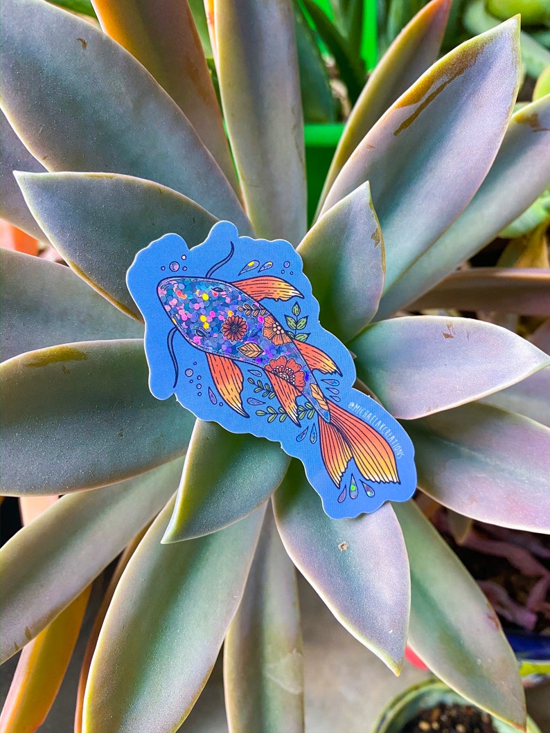 K&ouml;nnte beinhalten: Ein blauer und orangefarbener Fisch-Aufkleber mit Glitzer und floralen Details. Der Aufkleber befindet sich auf einer gr&uuml;nen Sukkulentenpflanze.