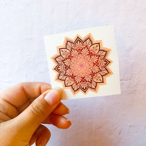 Pu&ograve; includere: Un adesivo con un design di mandala dettagliato in tonalit&agrave; arancio e marrone. Il mandala &egrave; circondato da un bordo bianco.