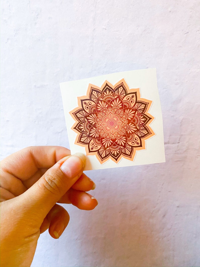 Pu&ograve; includere: Un adesivo con un design di mandala dettagliato in tonalit&agrave; arancio e marrone. Il mandala &egrave; circondato da un bordo bianco.