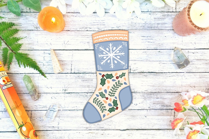 Puede incluir: Una media navide&ntilde;a azul y crema con un dise&ntilde;o de copo de nieve y hoja de acebo. La media est&aacute; sobre un fondo de madera blanco.